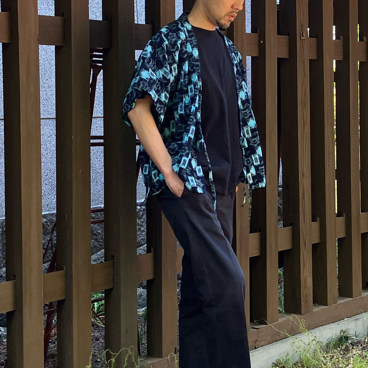 Side view of a man in a modern styled Noragi, leaning against a wooden fence.