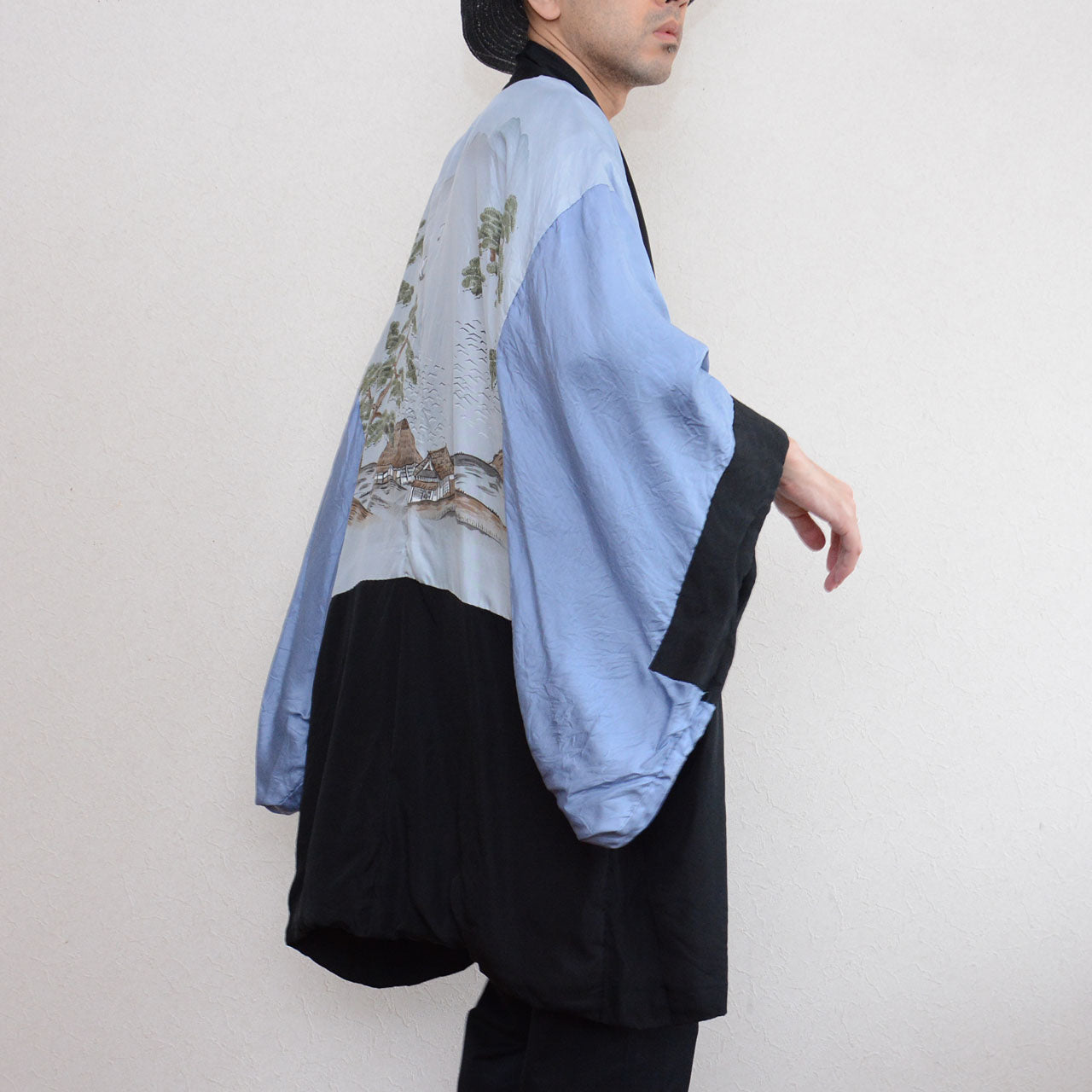 Black silk montsuki haori jacket worn inside out, side view showing pale blue silk landscape lining (haura) with hand-painted pine trees and thatched farmhouses, black outer fabric at lower hem and sleeve cuff, Japan vintage 1970s–80s