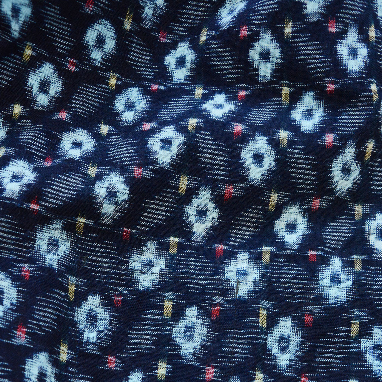 indigo kasuri cotton — close-up of ikat weave with light blue, red, and yellow accent threads on deep navy ground