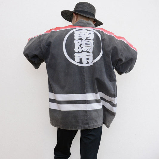 Model wearing a faded charcoal-gray Japanese firefighter happi jacket with a large circular kanji crest on the back