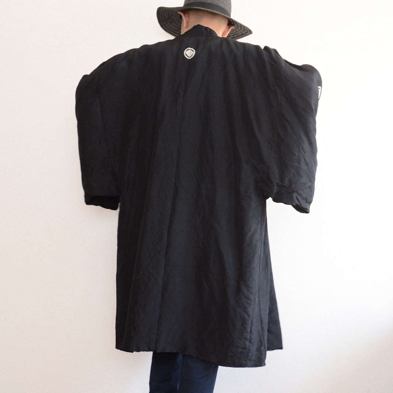 Model wearing men's black itsutsumon haori jacket, back view showing long silhouette and back family crest, styled with cap, model height 171 cm