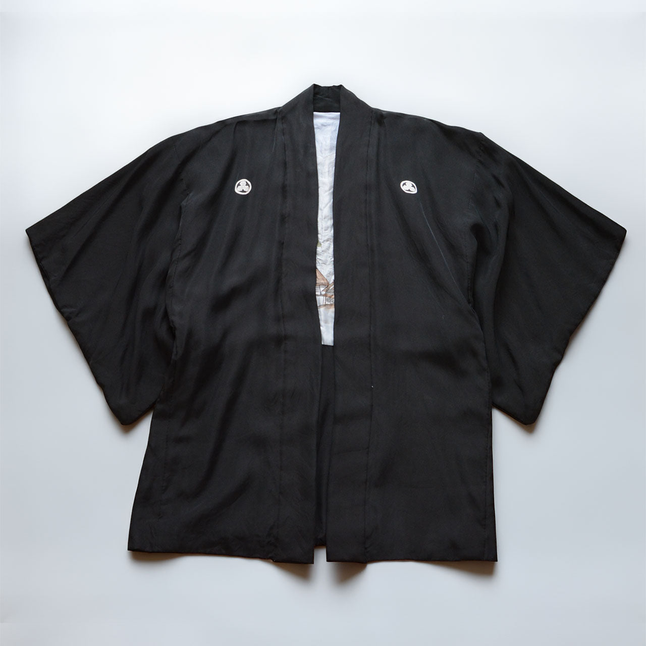 Black silk montsuki haori jacket, solid black outer fabric with two white Maru ni Mitsu Kashiwa family crests on chest panels, pale blue landscape lining visible at center front opening, Japan vintage 1970s–80s