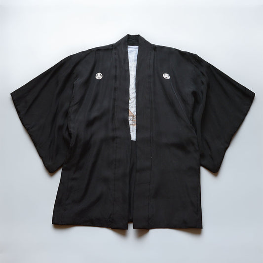 Black silk montsuki haori jacket, solid black outer fabric with two white Maru ni Mitsu Kashiwa family crests on chest panels, pale blue landscape lining visible at center front opening, Japan vintage 1970s–80s