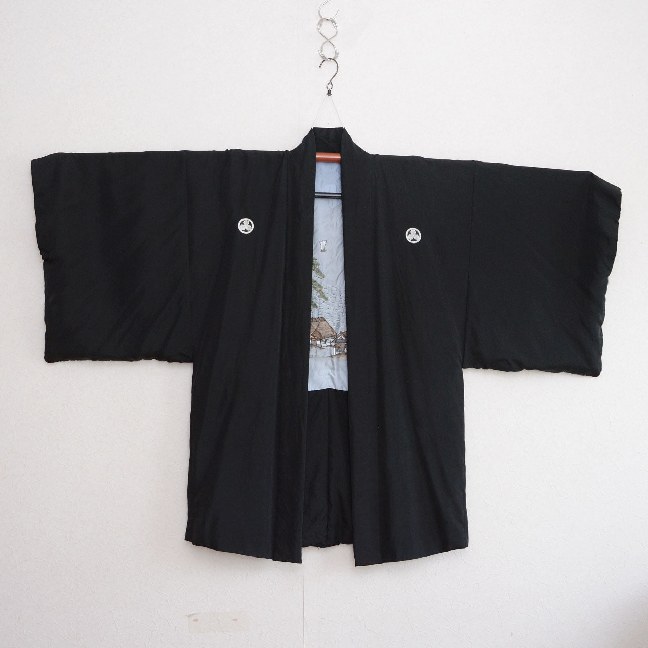 Black silk montsuki haori jacket on hanger, two white Maru ni Mitsu Kashiwa family crests on chest panels, pale blue hand-painted landscape lining with thatched farmhouse and pine trees visible at center front, Japan vintage 1970s–80s