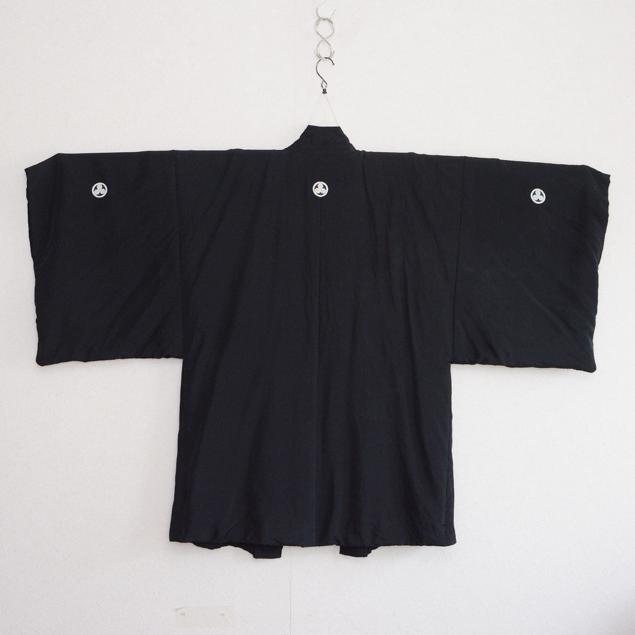 Black silk montsuki haori jacket on hanger, back view showing three white Maru ni Mitsu Kashiwa family crests — one on back center, one on each sleeve — solid black silk fabric, Japan vintage 1970s–80s
