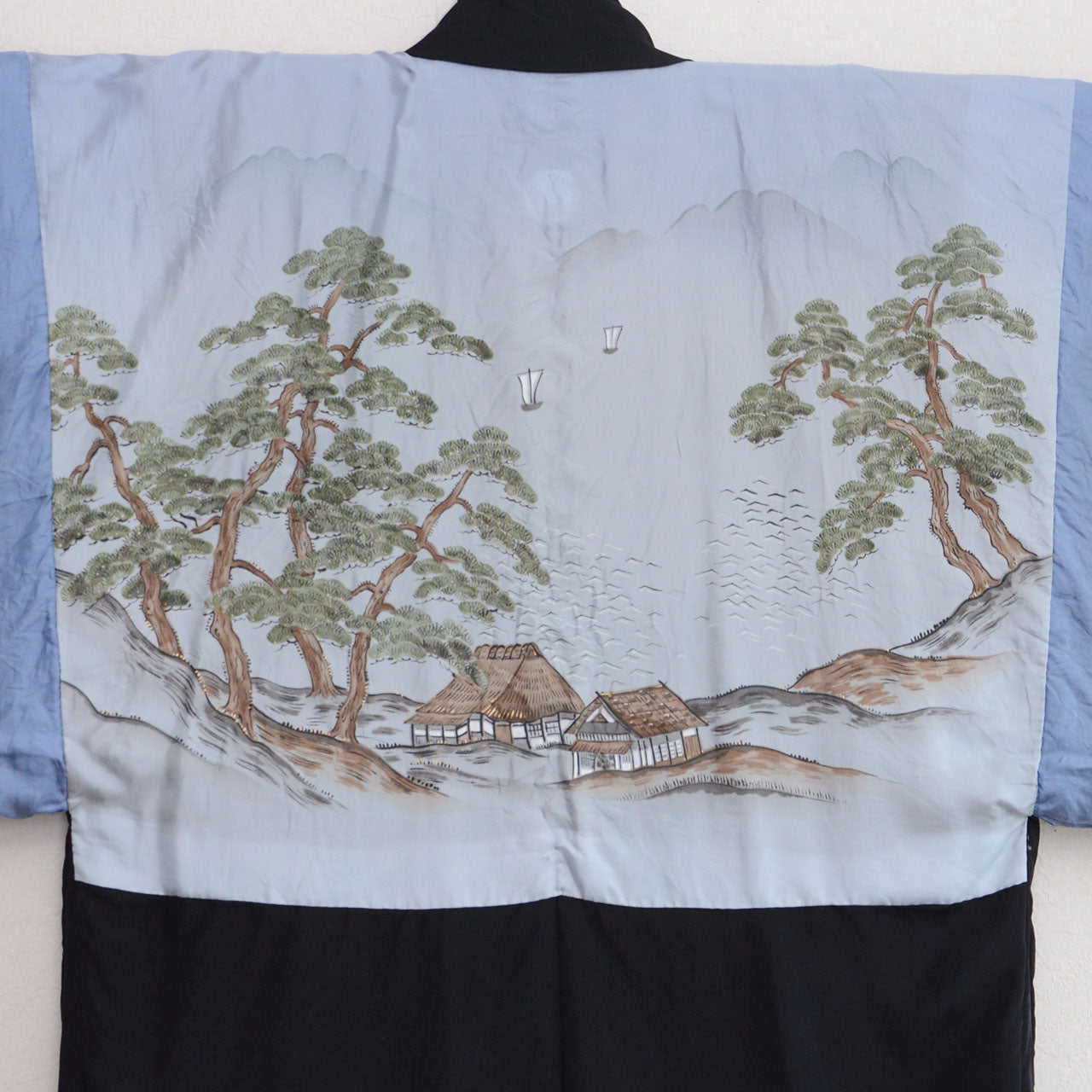 Black silk montsuki haori jacket, back view showing pale blue hand-painted landscape lining (haura) with pine trees, thatched farmhouse, sailing boat and mountains, black outer fabric visible at collar and lower hem, Japan vintage 1970s–80s