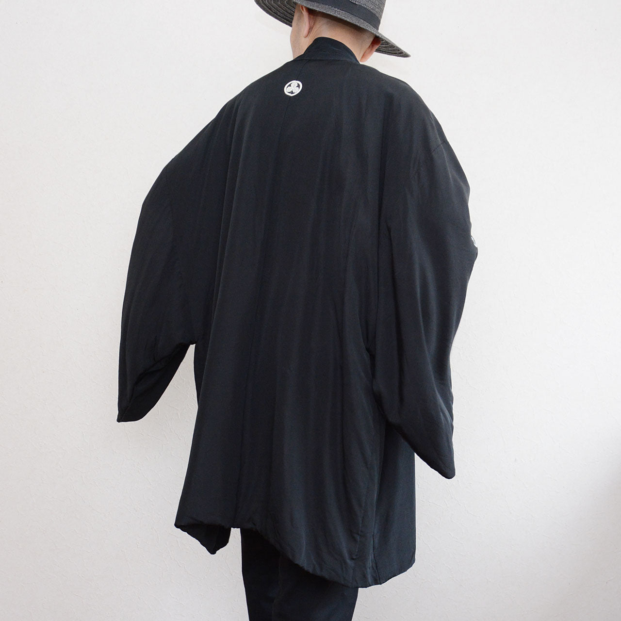 Black silk montsuki haori jacket, back view showing white Maru ni Mitsu Kashiwa family crest at upper back center, solid black silk outer fabric with wide sleeve silhouette, Japan vintage 1970s–80s