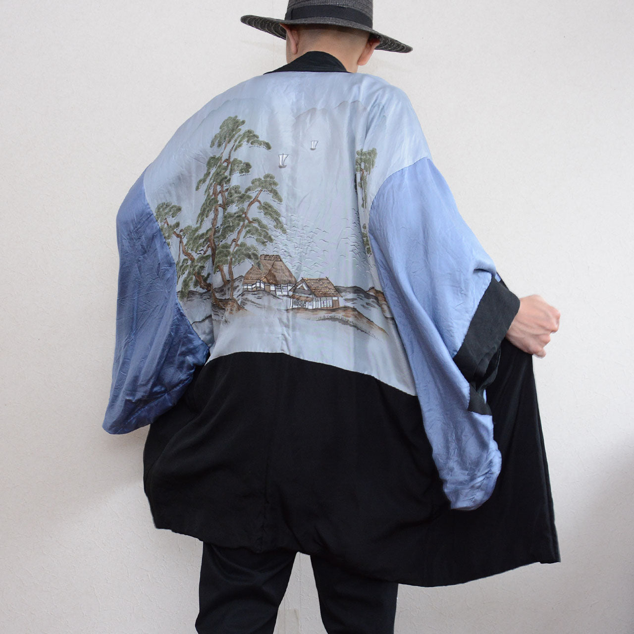 Black silk montsuki haori jacket worn inside out, back view showing full pale blue silk landscape lining (haura) with hand-painted pine trees, thatched farmhouses and water scene, black outer fabric visible at collar and lower hem, Japan vintage 1970s–80s