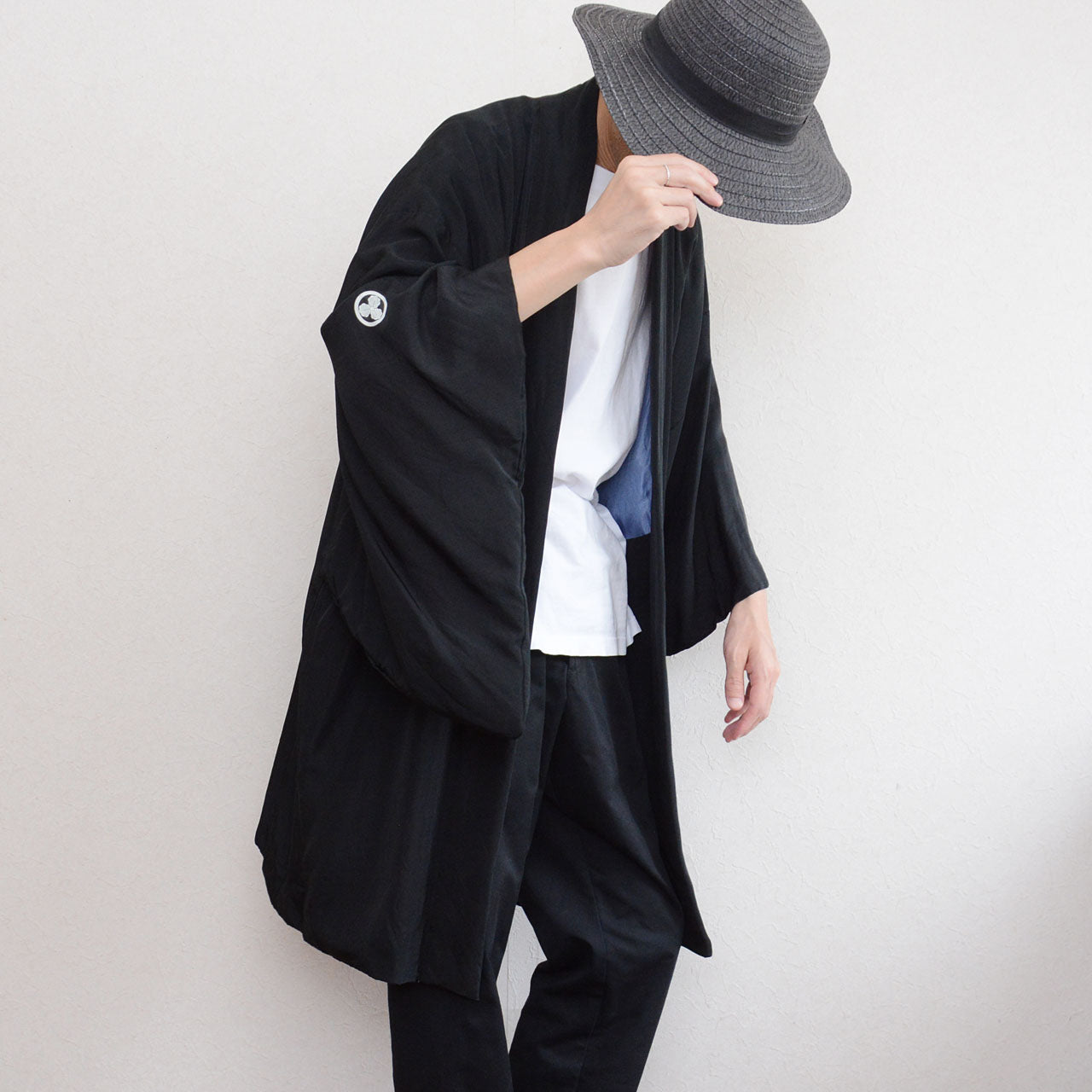 Black silk montsuki haori jacket worn open over white t-shirt and black trousers, three-quarter front view showing Maru ni Mitsu Kashiwa family crest on sleeve, pale blue landscape lining visible at opening, Japan vintage 1970s–80s