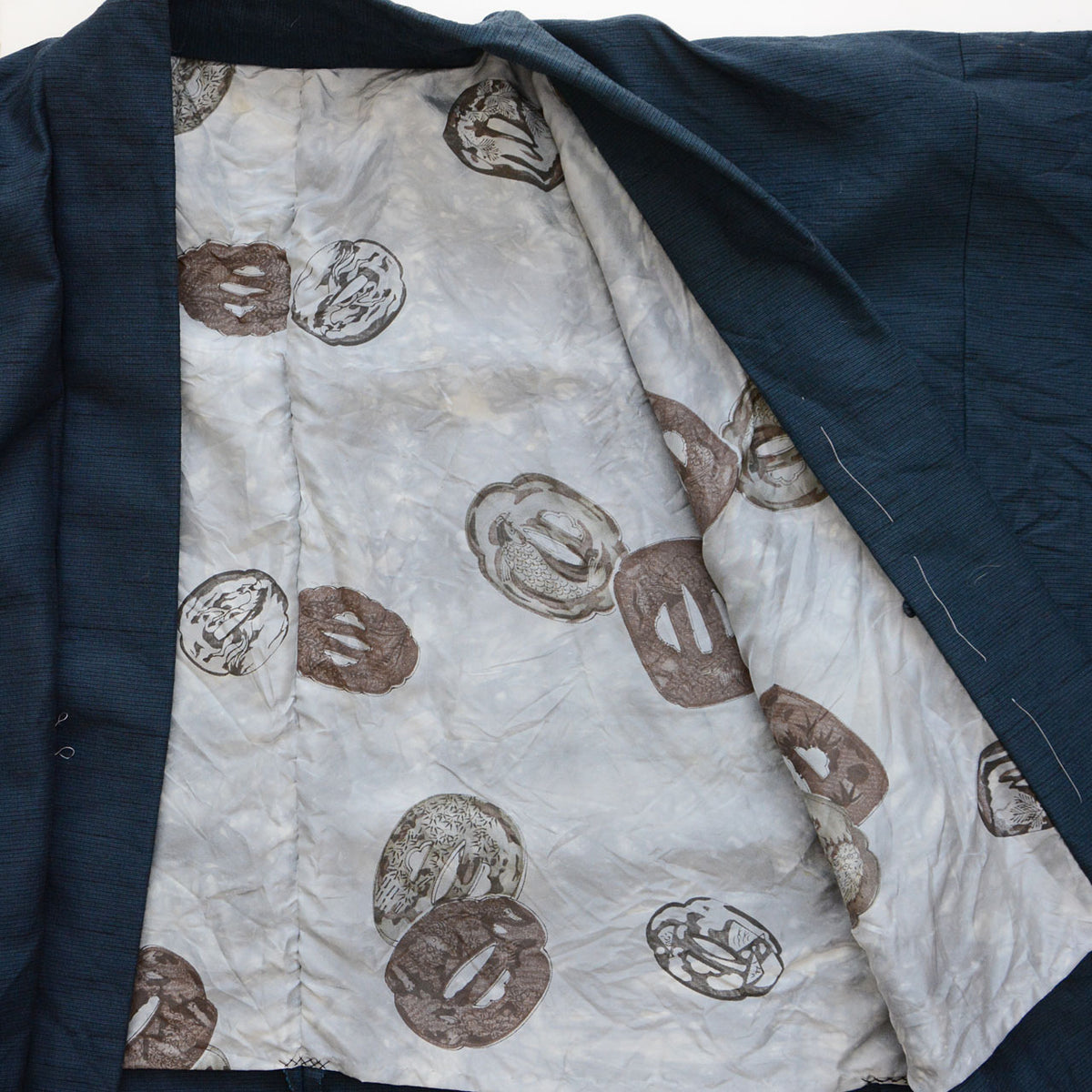 Light gray haori lining with scattered tsuba sword-guard motifs in brown and charcoal, contrasting with deep blue-green outer fabric.