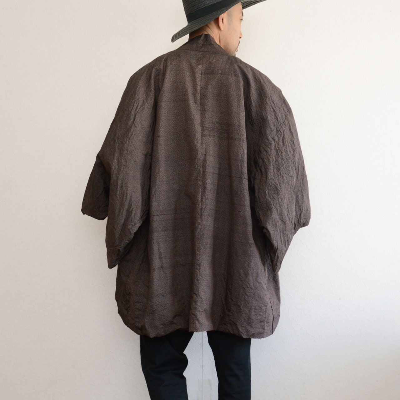 Man wearing a dark brown vintage Japanese haori jacket — back view, wide sleeve silhouette, layered over white t-shirt and black trousers, straw hat