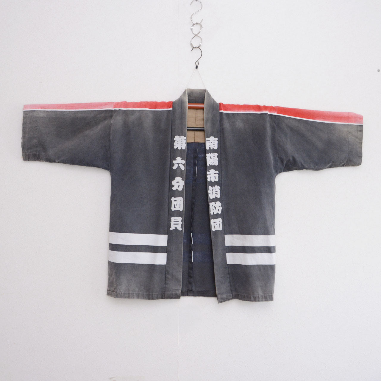 Front of a faded charcoal-gray cotton Japanese firefighter happi jacket hanging with its sleeves extended, featuring red and white shoulder stripes and two white horizontal stripes near the hem