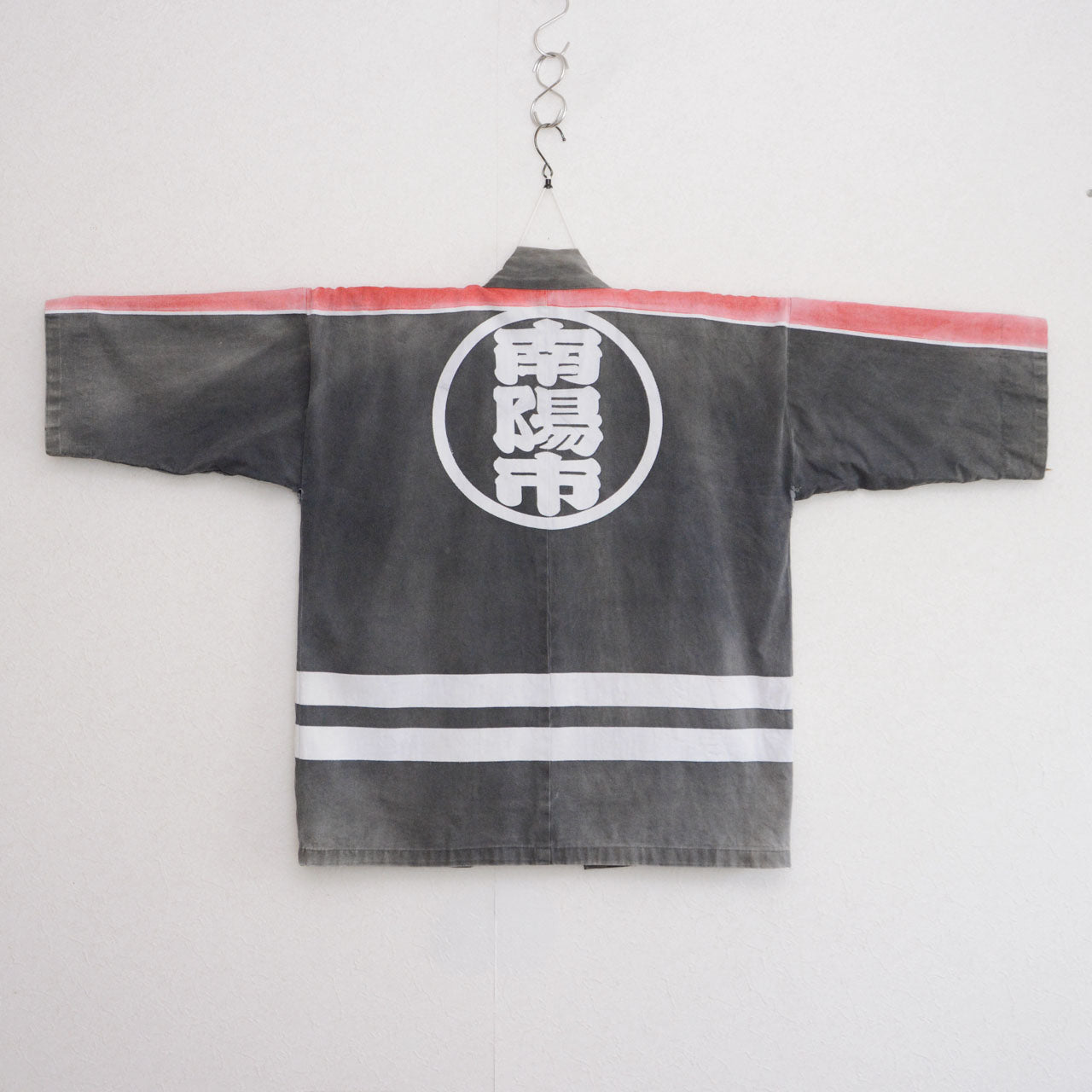 Back of a faded charcoal-gray cotton Japanese firefighter happi jacket with a large circular white design and two white horizontal stripes near the hem