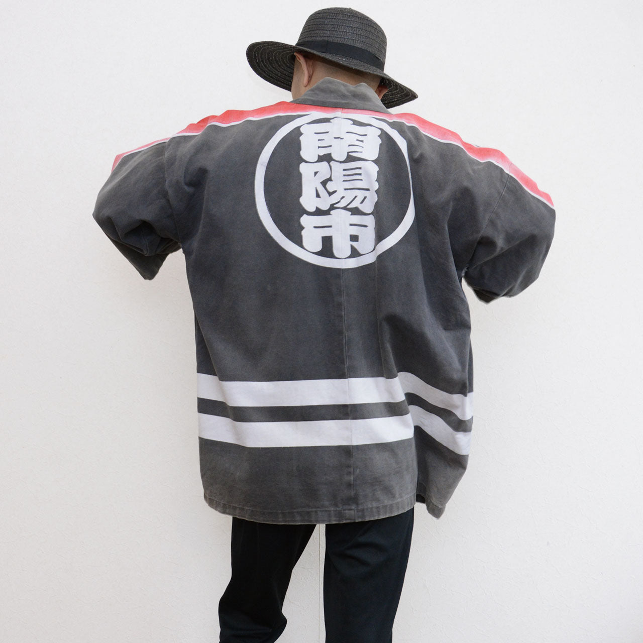 Model wearing a faded charcoal-gray Japanese firefighter happi jacket with a large circular kanji crest on the back