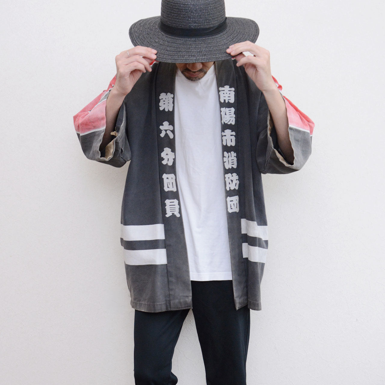 Model wearing a faded charcoal-gray Japanese firefighter happi jacket with white collar lettering and red and white shoulder stripes