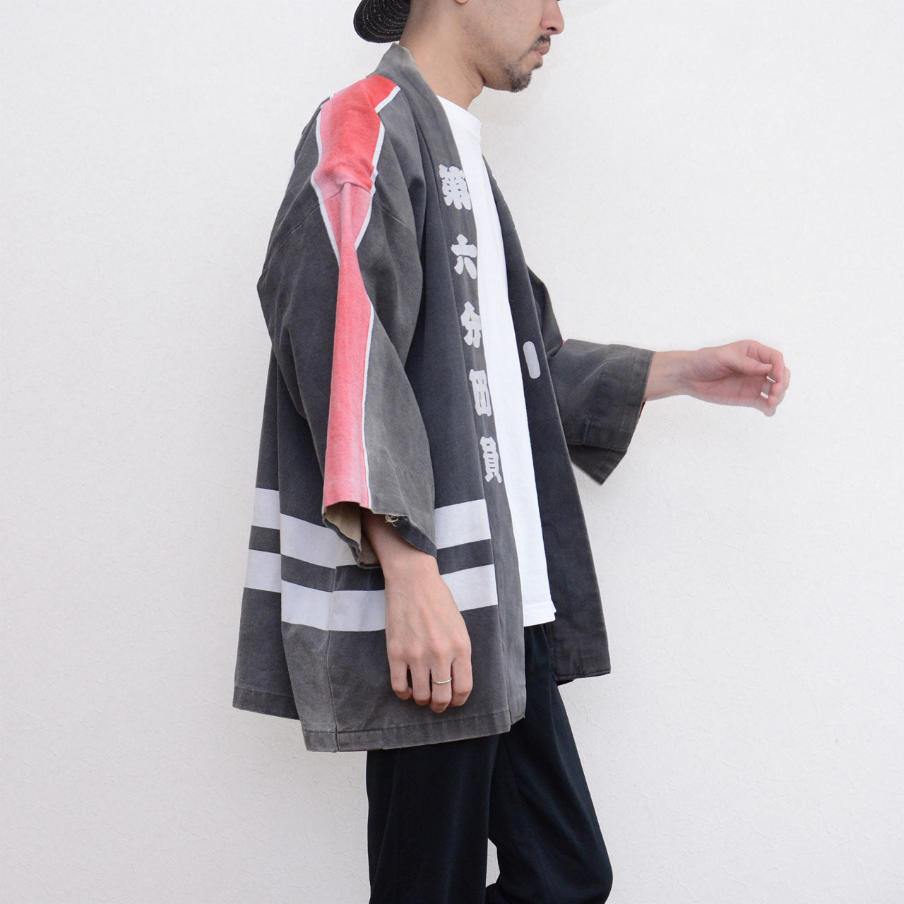 Side view of a model wearing a faded charcoal-gray Japanese firefighter happi jacket with red and white shoulder stripes and white horizontal stripes near the hem