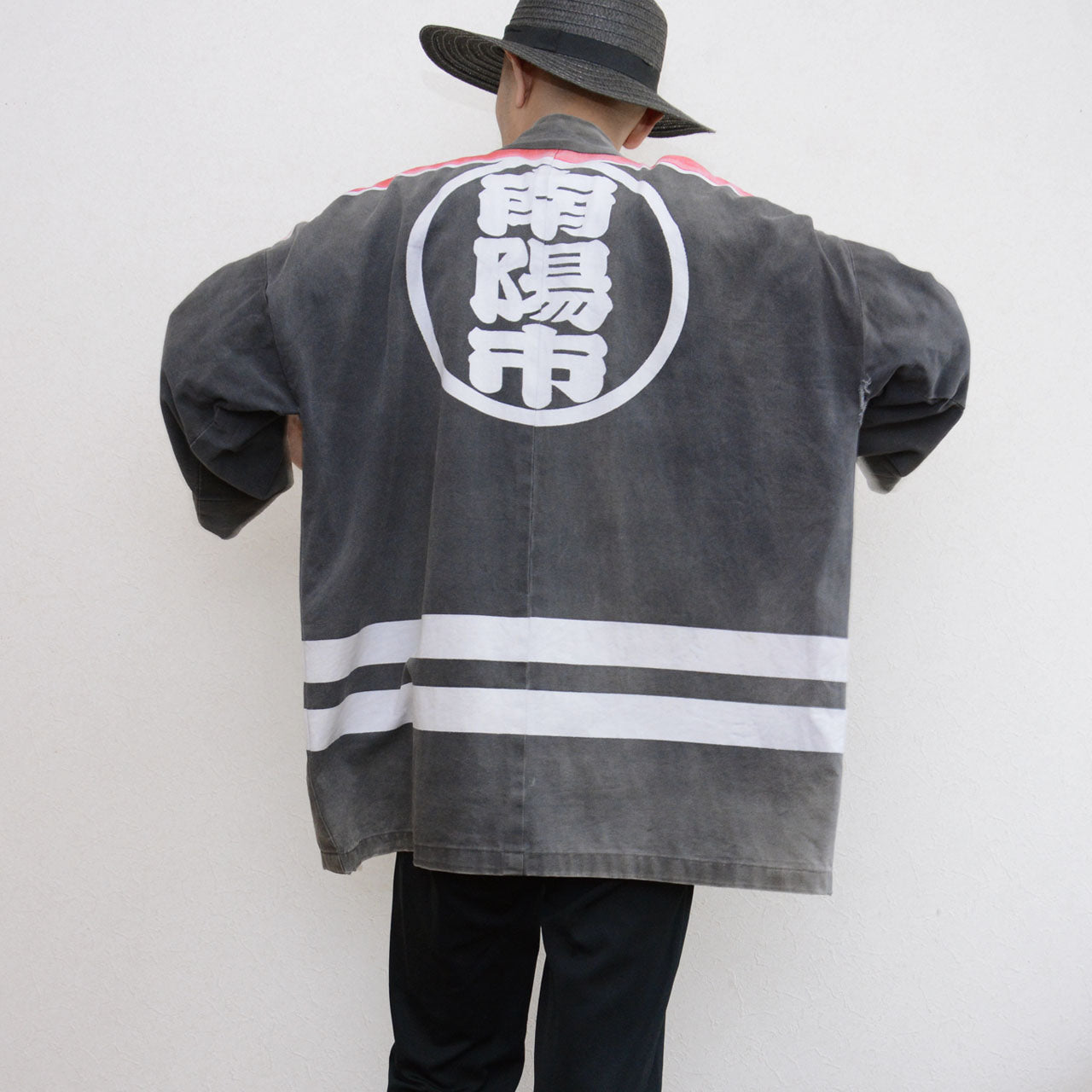 Model wearing a faded charcoal-gray Japanese firefighter happi jacket with a large circular kanji crest on the back