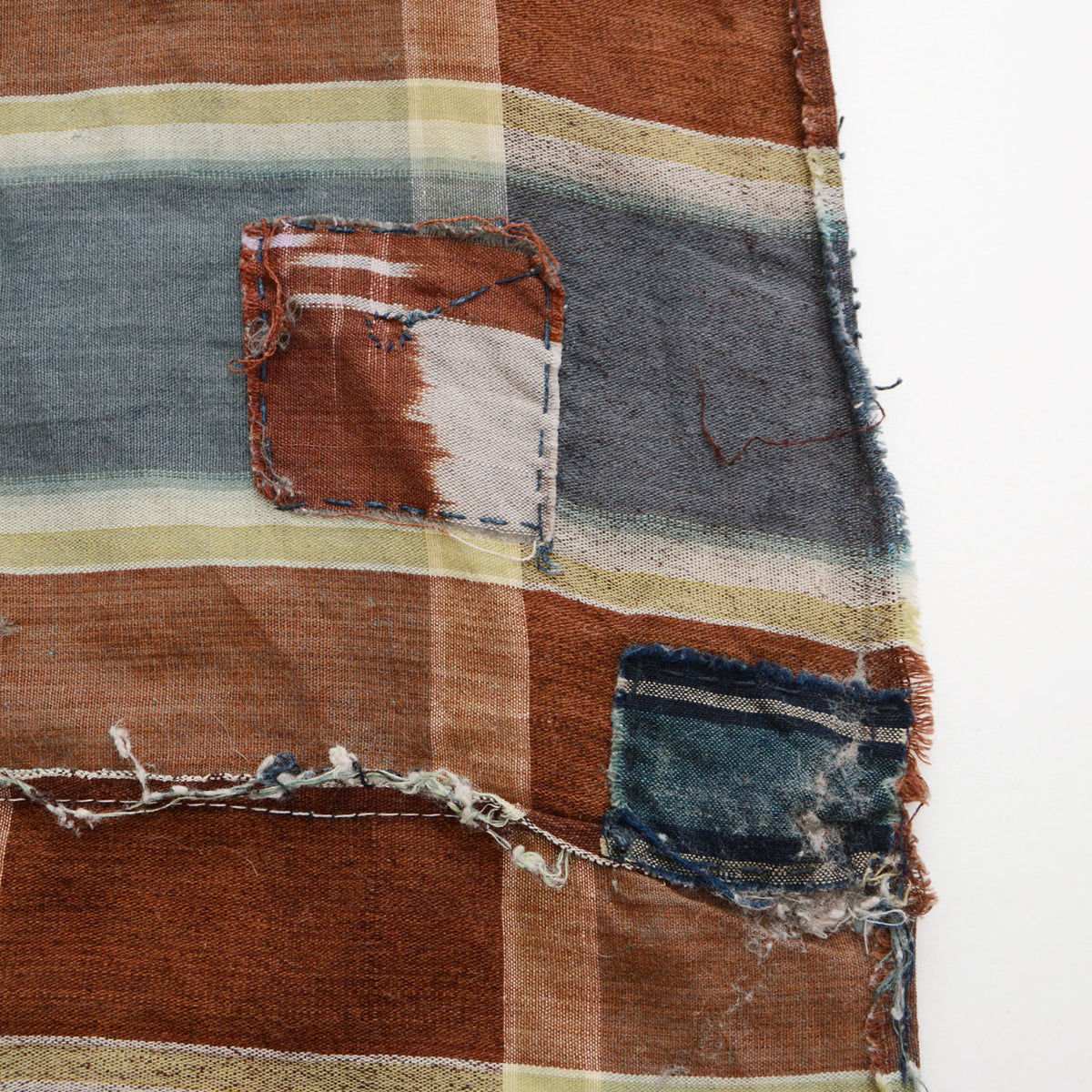 Brown, blue-gray, and cream checkered cotton fabric with multiple contrasting repair patches, frayed edges, and exposed threads